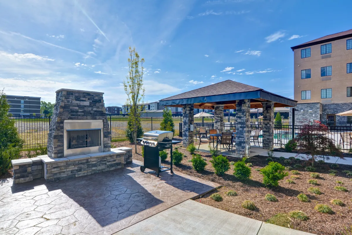An exterior view showcasing a covered pavilion with a stone fireplace, grill, and landscaped garden. The scene is set up for outdoor gatherings, dining, and relaxation, presenting a welcoming environment for unwinding after a productive day.