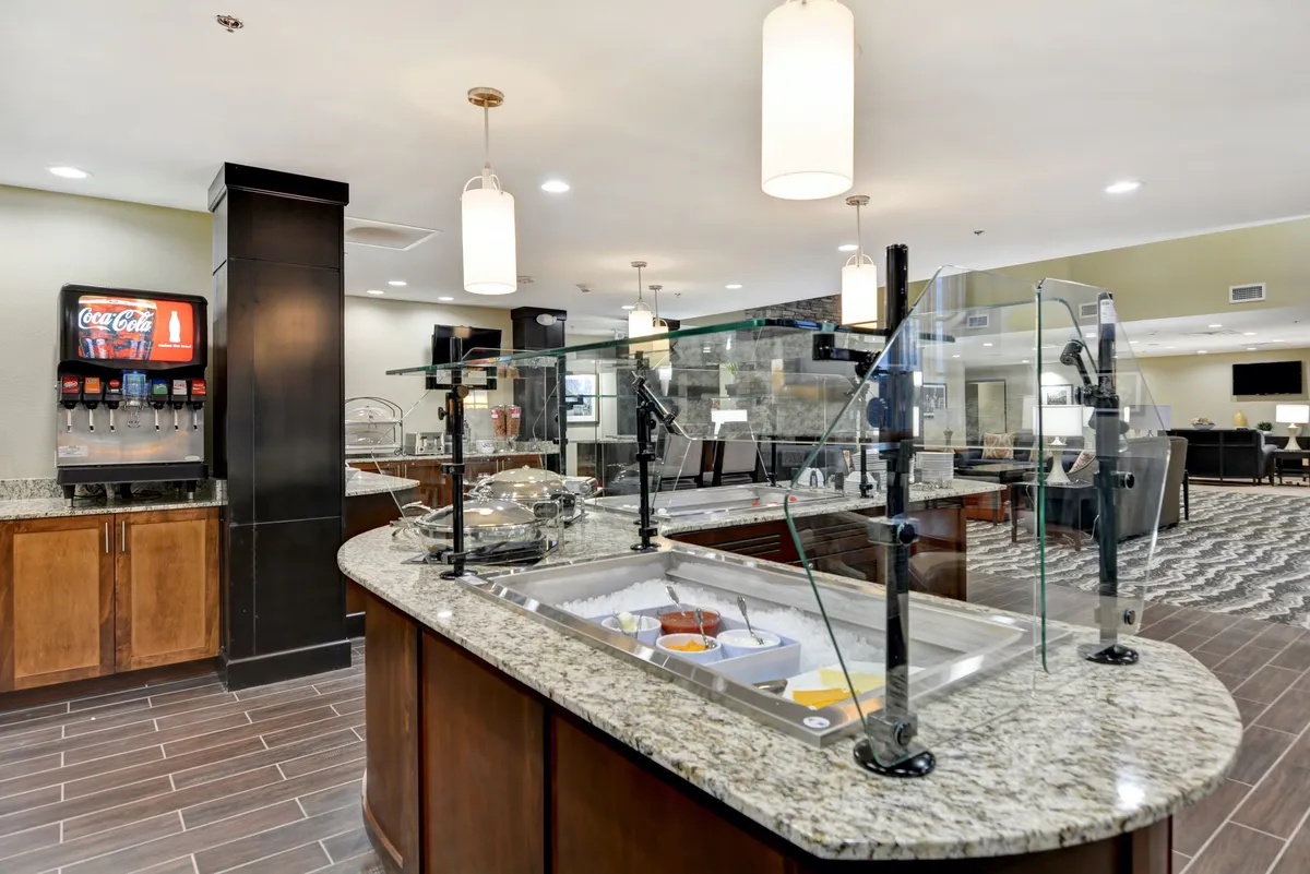 A bright, inviting breakfast area with a granite countertop serving space, a Coca-Cola dispenser, hanging light fixtures, and guests enjoying their morning meal. The mood is casual and welcoming, ideal for starting the day in comfort.