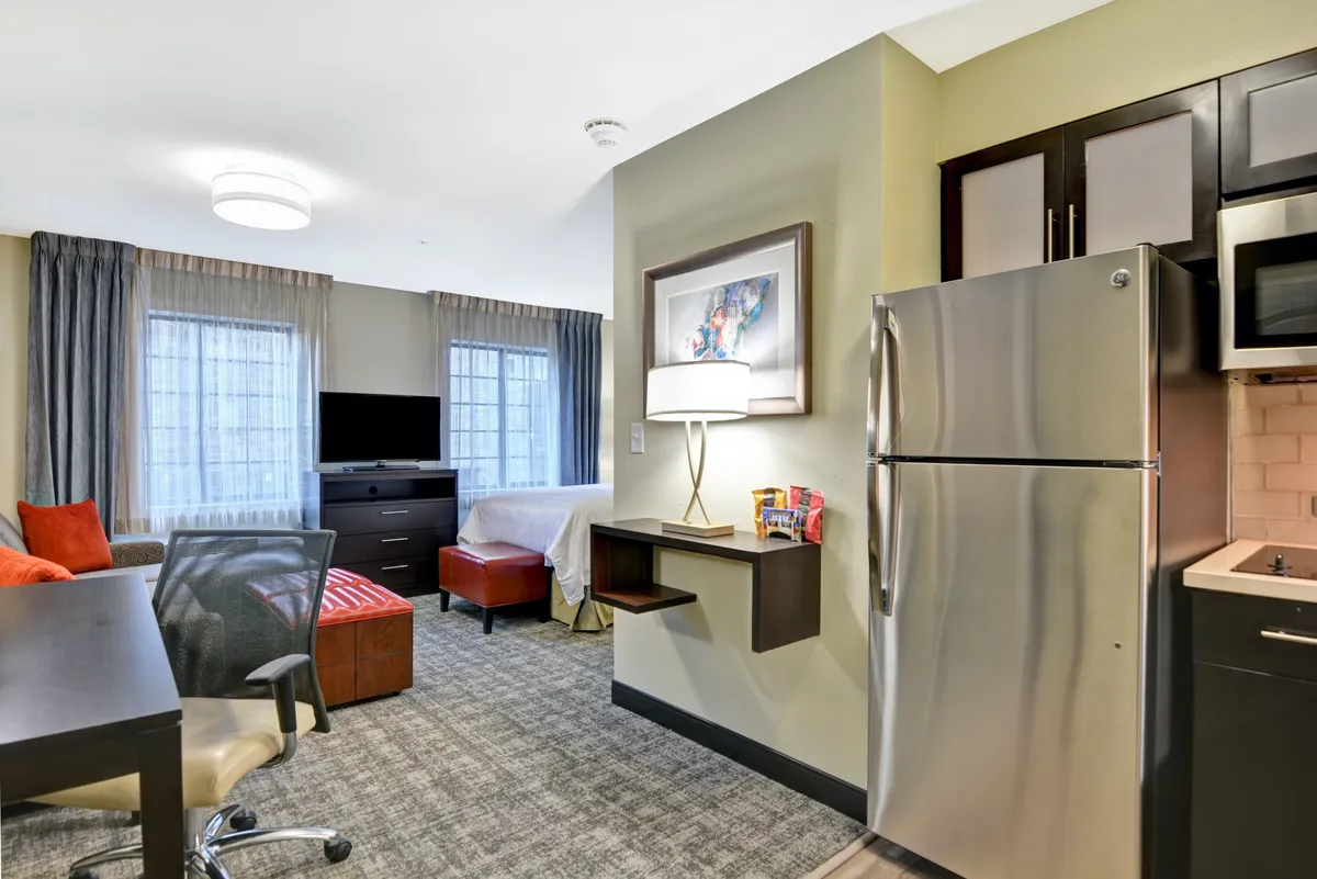 The image showcases a bright, modern studio interior featuring a comfy sofa, a plush bed, a well-equipped kitchen, and a dedicated workspace with a desk and chair, all arranged in an inviting, open-plan layout.