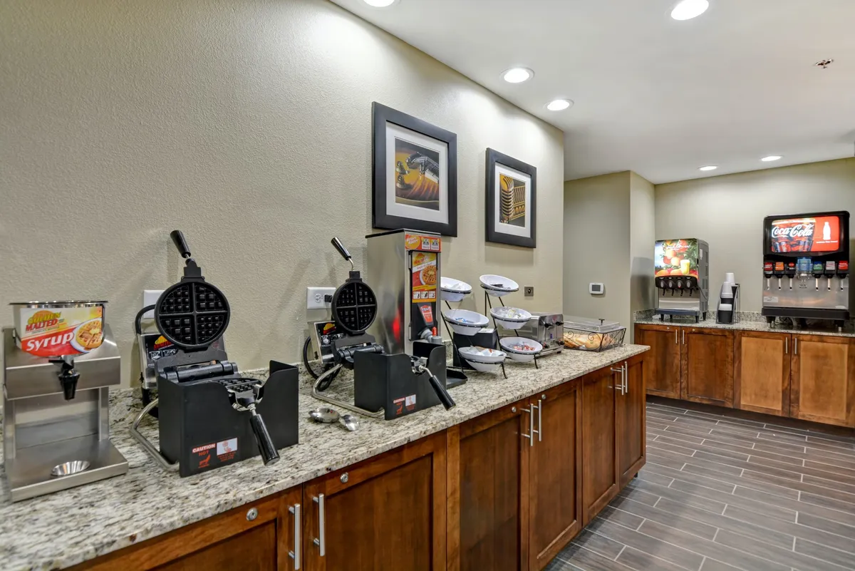 This photo shows a breakfast setup with waffle makers, a syrup dispenser, a beverage station, and decorative art frames. The scene conveys the availability of kitchen appliances and encourages a sense of homestyle meal preparation.