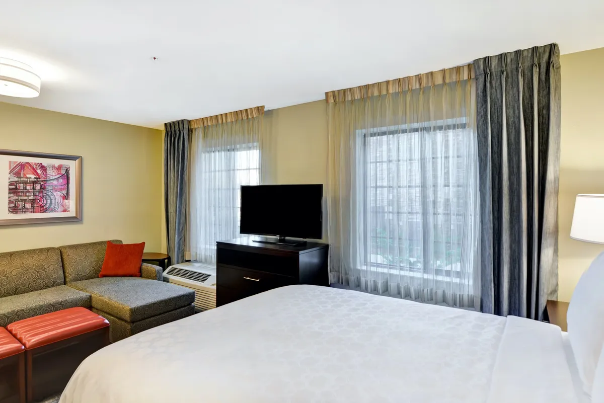 A bright and cozy studio interior featuring a plush queen bed, a comfortable sofa, a wall-mounted TV, large windows letting in natural light, and artful decorative touches. The layout appears open and accessible, with a blend of soft furnishings and personal décor.