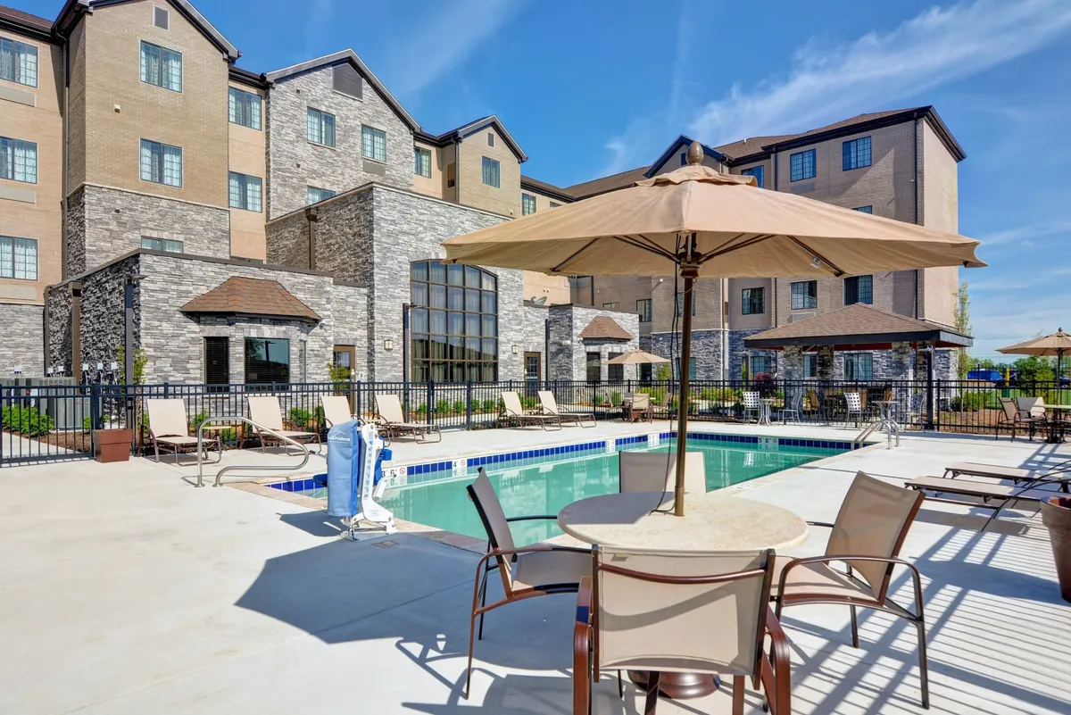 A vibrant outdoor pool scene featuring sparkling blue water, poolside lounge chairs, umbrellas for shade, and the building's facade in the background. The setting is sunny and cheerful, with guests relaxing and socializing by the water.