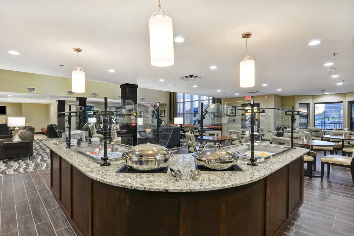 This image features a granite countertop with serving stations, hanging light fixtures above, and a visible adjacent seating area. The contemporary design and open space suggest a multi-functional kitchen or dining area, with room for meal preparation and casual dining.